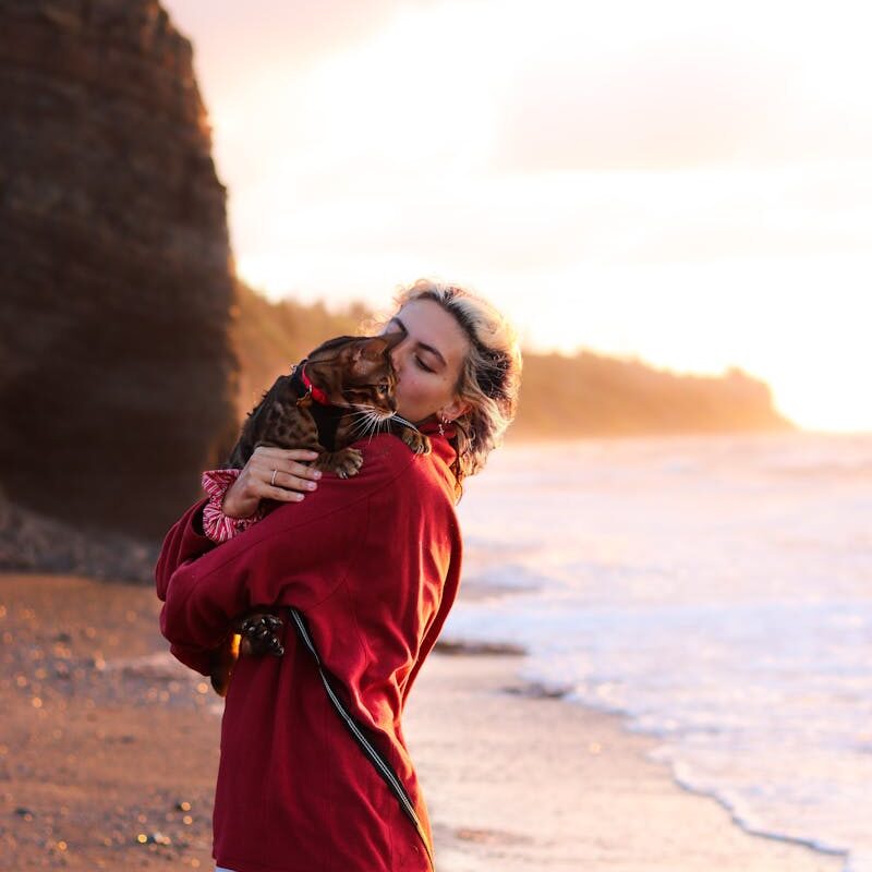 A woman lovingly holds her cat by a coastal cliff at sunset, showcasing serene companionship.