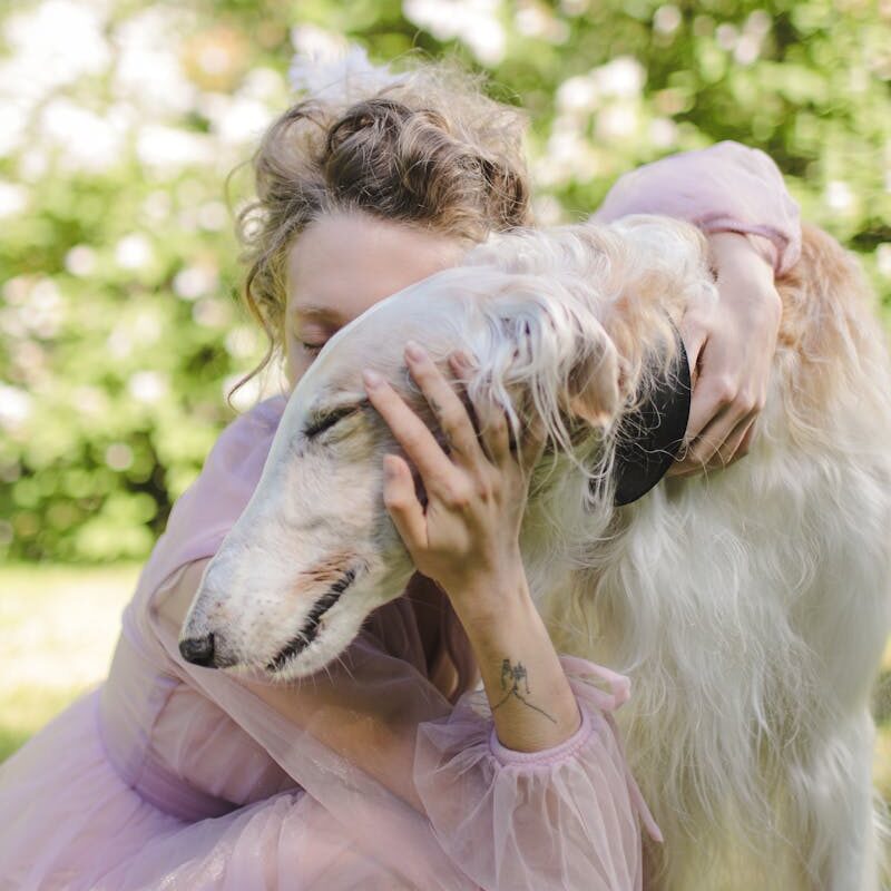 A woman lovingly embraces a Borzoi dog outdoors in a blooming garden setting.